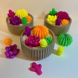 Puede incluir: Tres macetas de hormigón llenas de velas con forma de suculentas en colores vibrantes. Las velas son en tonos de amarillo, naranja, verde, rosa y morado. Las macetas están dispuestas sobre una superficie blanca.