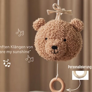 Può includere: Un mobile a forma di orsetto marrone con anello e fiocco in legno. Il mobile presenta il testo "you are my sunshine" e note musicali. Un anello di legno è mostrato con il nome "Emilia".