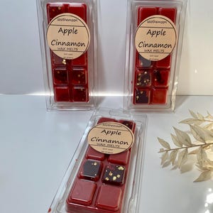 Könnte beinhalten: Drei transparente Kunststoffverpackungen mit roten Wachs-Schmelzstücken. Jede Verpackung hat ein rundes Etikett mit der Aufschrift "Apple Cinnamon WAX MELTS". Einige der Wachswürfel haben dunkelbraune und goldene Flecken.