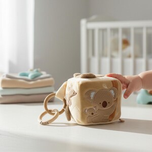 Könnte beinhalten: Beigefarbener Baby-Aktivitätswürfel mit Koala-Bär-Design. Der Würfel hat Holzringe und den eingravierten Namen "Emilia". Im Bild sind auch gefaltete Decken und ein Kinderbett zu sehen.