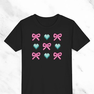 Puede incluir: Camiseta negra con un diseño pixelado de lazos rosas y corazones azules.