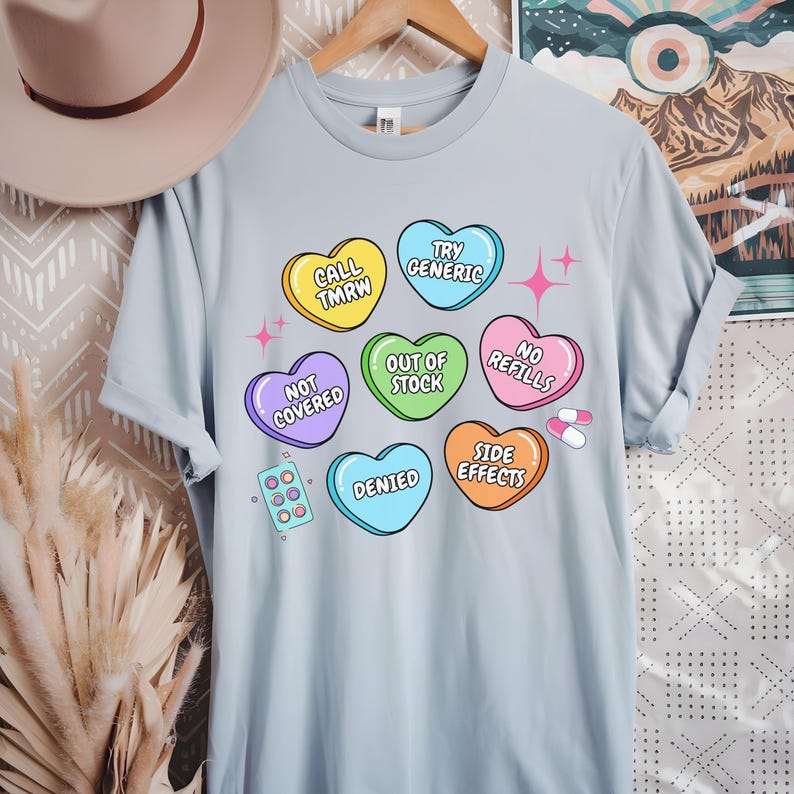 May include: Light blue t-shirt with a graphic of colorful candy hearts with phrases like "Call Tmrw", "Try Generic", "Out of Stock", "No Refills", "Side Effects", "Denied", and "Not Covered".