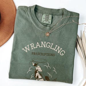 May include: Sage green t-shirt with the words "WRANGLING PRESCRIPTIONS" in a vintage font. The shirt features a cowboy roping a horse graphic. A brown hat and a silver necklace with a pendant are also visible.