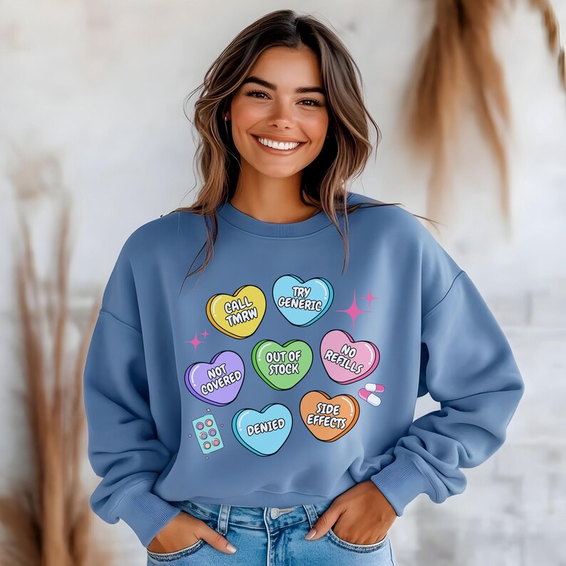 May include: A blue sweatshirt with a graphic of colorful candy hearts that say "Call Tmrw", "Try Generic", "Out of Stock", "No Refills", "Not Covered", "Denied", and "Side Effects".