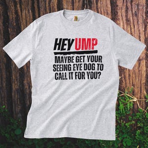 Könnte beinhalten: Ein graues T-Shirt mit dem Text "HEY UMP MAYBE GET YOUR SEEING EYE DOG TO CALL IT FOR YOU?" in Schwarz und Rot gedruckt.