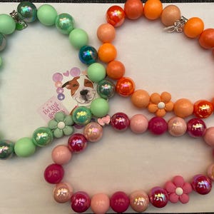 May include: Three colorful beaded bracelets with flower charms. The bracelets are green, orange, and pink.