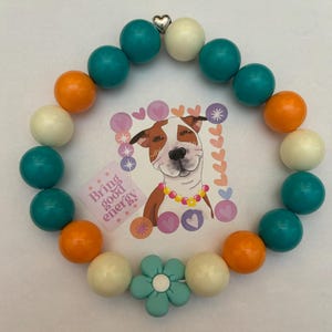 May include: A teal, white, and orange beaded bracelet with a turquoise flower charm. The bracelet has a silver heart charm and the text "Bring good energy!" is visible in the background.