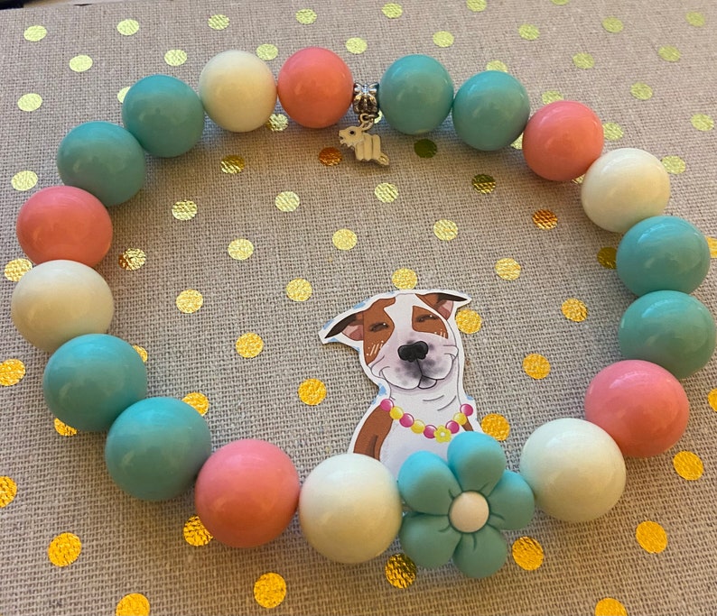 May include: A pastel-colored beaded bracelet with a silver charm in the shape of a dog. The beads are white, light blue, and pink.
