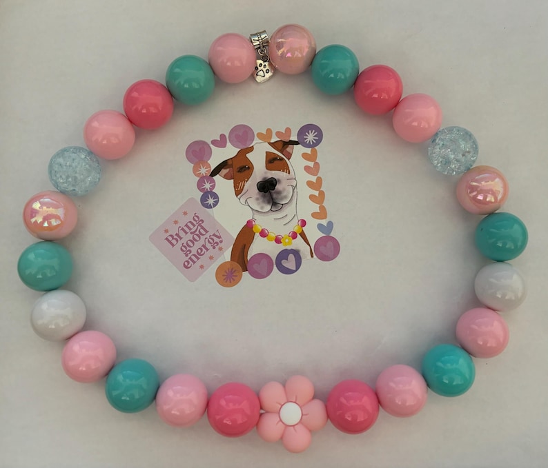 May include: A colorful beaded necklace with pink, blue, and white beads. The necklace has a flower charm and a silver clasp. The necklace is on a white background.  "Bring good energy" is written on a sticker in the background.
