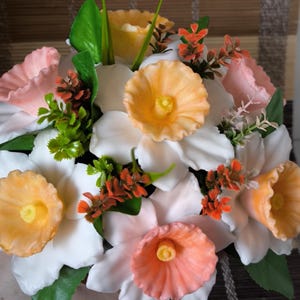 May include: A vibrant floral display featuring white, peach, and orange flowers. The arrangement includes green leaves and small orange and green accent flowers. The flowers have a detailed, textured appearance, suggesting they may be handmade.