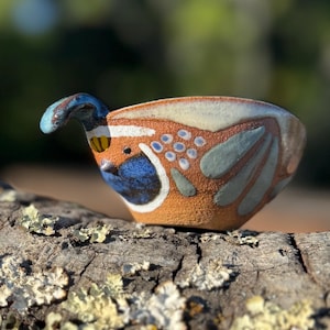 May include: A handcrafted ceramic bird-shaped bowl with a handle. The bowl features a brown base with blue, white, and green painted details. The handle is blue and the bowl is resting on a piece of wood.