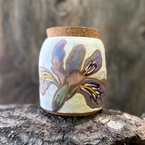 May include: A cylindrical ceramic jar with a textured off-white surface featuring a hand-painted purple and yellow iris flower design. The container is topped with a round, light brown cork lid that fits securely into the top opening of the vessel.