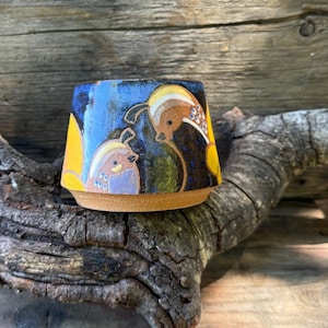 May include: A ceramic cup with a tapered shape and a natural clay base. The exterior features a deep blue glaze decorated with hand-painted illustrations of two quail birds in shades of yellow, brown, and white, set against a dark blue background.