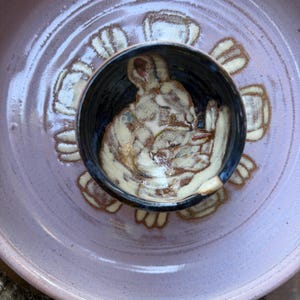 May include: A handmade ceramic bowl with a dark blue interior, featuring a detailed illustration of a rabbit. The bowl sits within a larger, lavender-colored plate with a floral design. The rabbit illustration is in shades of brown and cream.