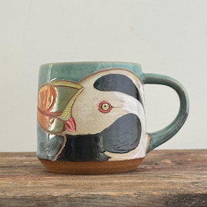 May include: A ceramic mug with a teal glaze and a hand-painted puffin illustration. The bird features a black and white head, a bright orange and yellow beak, and a small red eye. The base of the mug is unglazed brown clay.