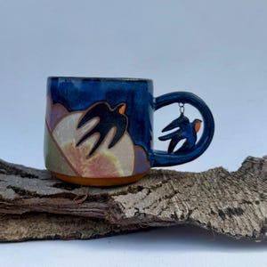 May include: A handmade ceramic mug with a dark blue glaze. The mug features a unique handle with a bird design and a landscape scene with birds. The mug is resting on a piece of wood.