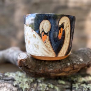 May include: A ceramic mug featuring a hand-painted design of two white swans with orange beaks against a deep blue background. The mug has a cylindrical shape with a smooth, glazed finish and a visible terracotta-colored base at the bottom.