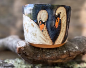 May include: A ceramic mug featuring a hand-painted design of two white swans with orange beaks against a deep blue background. The mug has a cylindrical shape with a smooth, glazed finish and a visible terracotta-colored base at the bottom.