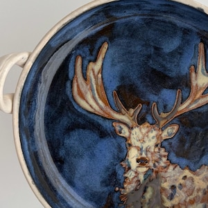 May include: A handmade ceramic bowl with a blue and brown glaze. The bowl features a detailed image of a deer with large antlers. The rim and handles are a cream color, creating a contrast with the darker interior.