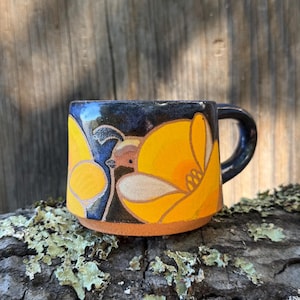 May include: A ceramic mug with a dark blue glazed upper section and a natural terracotta base. The mug features a hand-painted design of a quail surrounded by large, vibrant yellow poppy flowers with defined outlines.