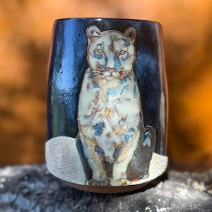 May include: A ceramic tumbler with a glossy dark blue and black glaze featuring a hand-painted white and tan mountain lion centered on the front. The base of the vessel has a textured, matte off-white finish, and the rim is smooth and rounded.
