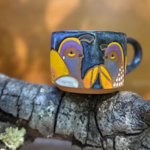 May include: A small, handmade ceramic mug with a dark blue glaze. The mug features a colorful, whimsical design of two birds with yellow, purple, and orange accents. The mug has a small handle and a brown base. The mug is sitting on a piece of wood.