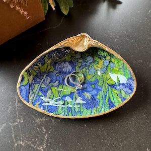 May include: A gold-rimmed seashell ring dish with a blue and green floral print. Two silver rings are inside the dish.
