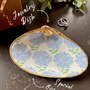 Puede incluir: Un plato de joyería de cerámica blanca con forma de concha, con un borde dorado y un patrón floral azul. El plato está sobre una superficie marrón oscura con un par de anillos de plata en la parte superior.