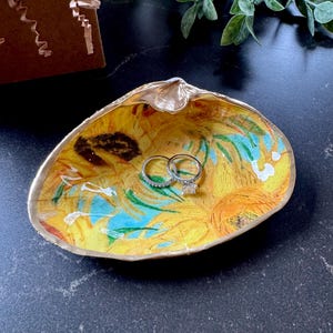 Puede incluir: Un joyero de cerámica con forma de concha con un diseño pintado de girasoles al estilo de Vincent van Gogh. Dos anillos de plata descansan en el joyero.