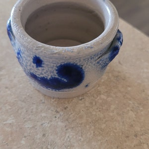 Può includere: Un piccolo vaso in ceramica bianca con smalto blu. Il vaso ha due piccole maniglie sui lati e un design maculato blu e bianco.
