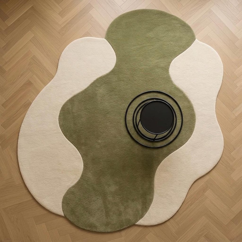 May include: An abstract area rug with a unique, flowing design. The rug features a combination of olive green and cream colors, with a black circular tray on top. The rug is placed on a light brown wooden floor.