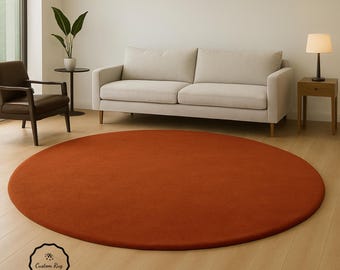 May include: A large, round, rust-colored rug dominates the foreground of a living room. The rug is placed on a light-colored wood floor, with a beige sofa and a dark brown armchair in the background. A small table with a lamp is also visible.