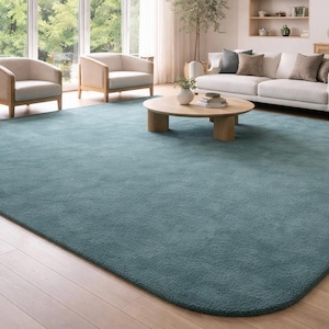 May include: A large, rectangular area rug with rounded corners, featuring a solid, deep teal blue colour and a soft, plush, textured pile surface. The rug is positioned flat on a light-toned wooden floor, covering a significant portion of the room.