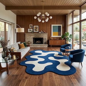 May include: A living room with a mid-century modern design, featuring a blue and white abstract rug. There's a grey sofa, two blue armchairs, a fireplace, wooden paneling, and a large window overlooking a patio.