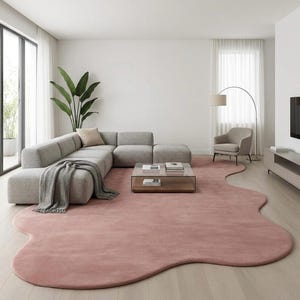 Puede incluir: Salón moderno con un gran sofá seccional gris claro, una mesa de centro con tapa de cristal y una alfombra rosa rubor de forma orgánica. Una manta gris y una lámpara de pie completan el diseño minimalista.
