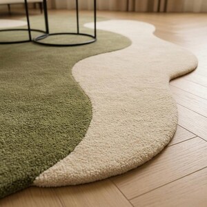 May include: A modern, abstract rug with a wavy design. The rug features two distinct sections: one in a muted green and the other in a cream color. The rug is placed on a wooden floor, and the design adds a touch of contemporary style to the space.