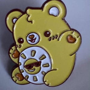 May include: A yellow bear-shaped enamel pin with a white sun design. The sun has a smiling face wearing sunglasses. The pin has brown outlines and details, including heart-shaped accents on the paws.