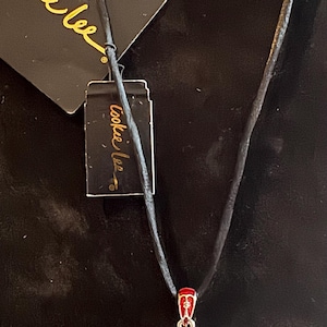 Pode incluir: Um colar com pingente vermelho e dourado com um cordão preto. O pingente tem uma moldura quadrada com o texto "Insert Photo Here". Uma etiqueta com o texto "Cookee Lee" está anexada.