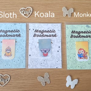 May include: Three magnetic bookmarks featuring different animal designs. The bookmarks are displayed on a wooden surface with decorative hearts and butterflies. The bookmarks depict a sloth, a koala, and a monkey, each holding a book. The text "Magnetic Bookmark" is printed on each bookmark.