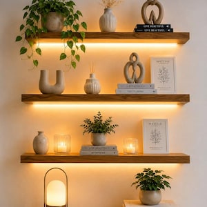 Op de afbeelding: Drie horizontale houten zwevende planken met geïntegreerde warme LED-verlichting, waarop diverse decoratieve items staan zoals potplanten, keramische vazen, abstracte sculpturen, kaarsen en boeken met titels zoals Live Beautiful en Matisse.