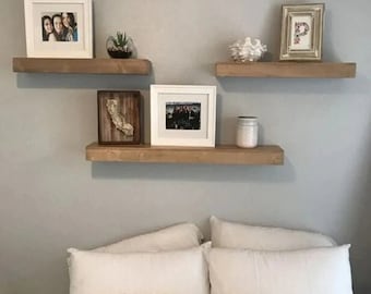 May include: Three wooden shelves mounted on a light gray wall with framed photos, a small potted plant, and decorative items on the shelves. A white bed with white pillows and a white throw blanket is in the foreground.