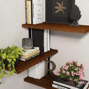 May include: A set of three wooden shelves with black metal brackets. The shelves are filled with books, a framed print, a small plant, and decorative objects. The shelves are mounted on a white wall.