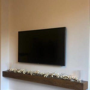 Può includere: Un grande televisore a schermo piatto nero montato su una parete beige. Sotto la TV c'è una mensola in legno marrone scuro decorata con una ghirlanda di bacche bianche e un portacandele in vetro trasparente. La stanza ha un'estetica moderna.