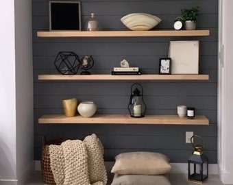 May include: Four wooden shelves mounted on a dark gray wall with various decorative items, including a small plant, a framed photo, a bowl, a globe, a geometric sculpture, a lantern, and candles. The shelves are made of light wood and have a natural finish.