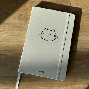Può includere: Un quaderno grigio chiaro con un'illustrazione di un gatto nero e la parola "toffied" stampata sulla copertina.