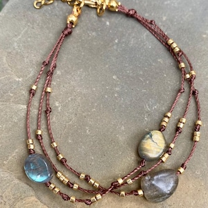 Peut inclure: Bracelet à plusieurs rangs avec trois brins de cordon marron, des perles dorées et trois pierres précieuses ovales polies bleues, jaunes et grises. Le bracelet est doté d'un fermoir doré.