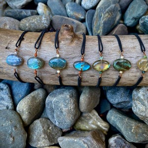 Peut inclure: Sept colliers avec des pierres précieuses ovales et irisées dans les tons de bleu, vert et jaune sont présentés sur du bois flotté. Chaque collier a un cordon noir et des accents cuivrés. L'arrière-plan présente divers rochers gris et bleus. Les pierres mesurent environ 2-3 cm.