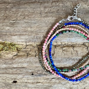 Peut inclure: Plusieurs bracelets de perles de différentes couleurs, dont rose, bleu, vert et or. Les bracelets sont composés de petites perles rondes et sont disposés en couches. Le fond est une surface en bois rustique.