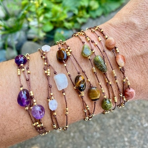 May include: A selection of gemstone bracelets in various colours and shapes. The bracelets are strung on brown cord with gold-coloured beads. Gemstones include amethyst, tiger's eye, and other natural stones.