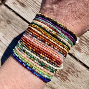 Könnte beinhalten: Eine Sammlung von Armbändern mit Perlen in verschiedenen Farben, darunter Blau, Grün, Rosa, Orange und Lila. Die Armbänder bestehen aus kleinen, runden Perlen und sind an einem Handgelenk gestapelt. Der Hintergrund ist eine Holzoberfläche.